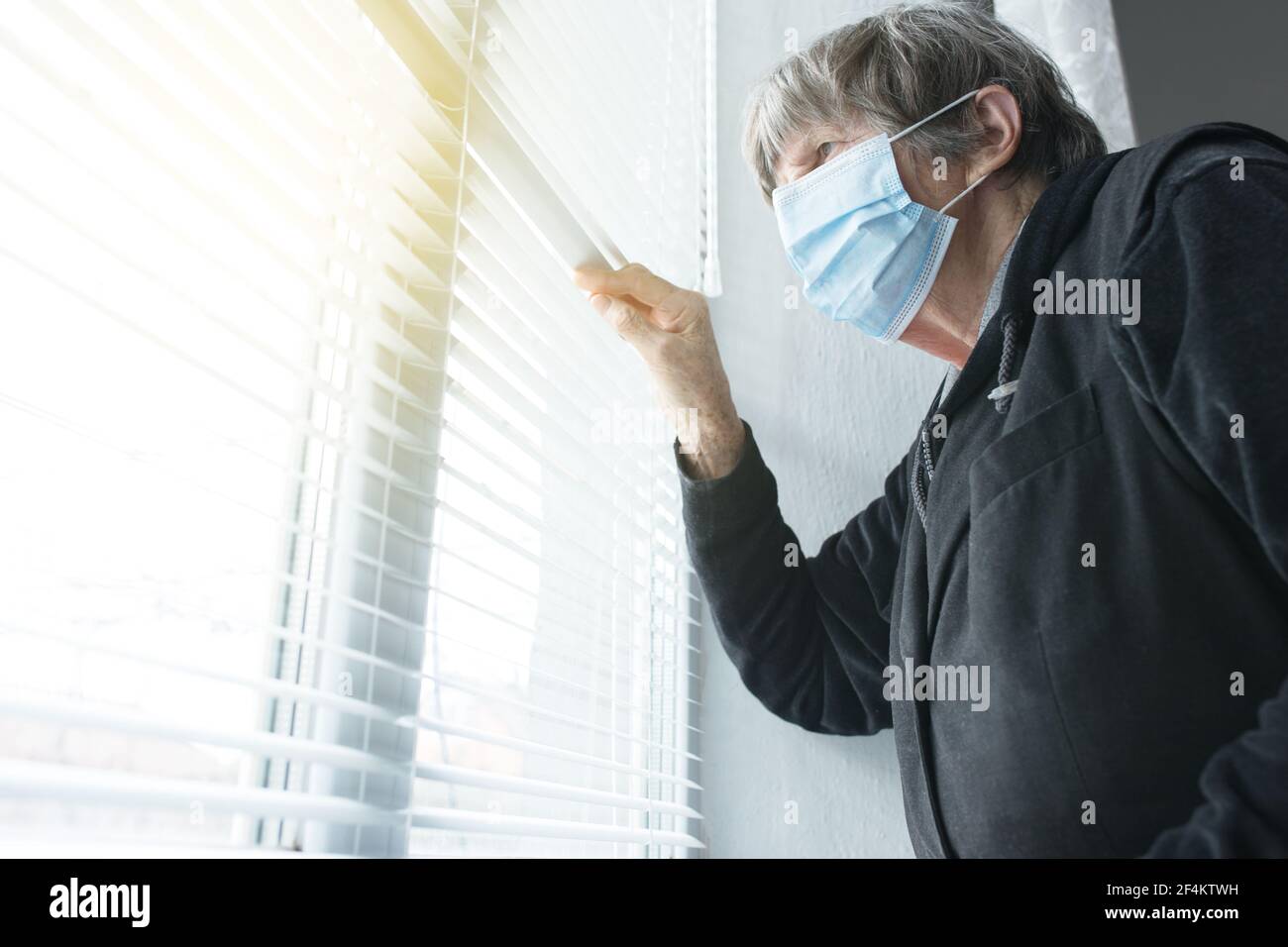 Old woman at home on self-isolation, in medical mask near the window looking outsid.e Stock Photo