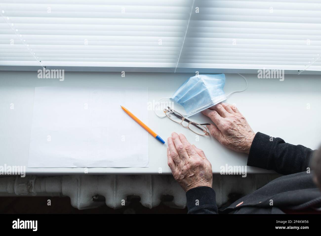 Hands of elderly woman writing a will at windowsill with medical mask and glasses. Stock Photo