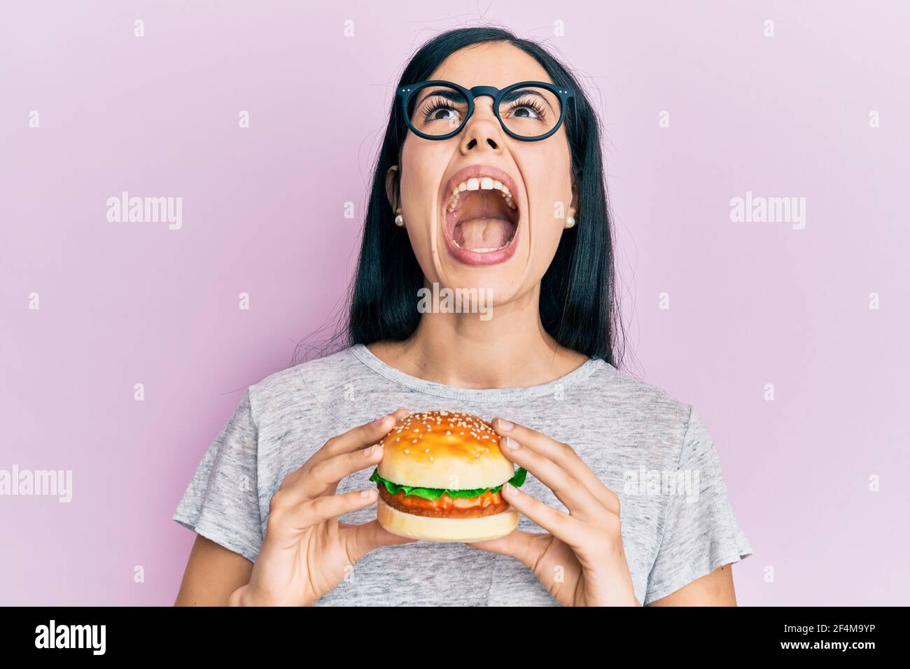 Beautiful young woman eating tasty hamburger angry and mad screaming ...