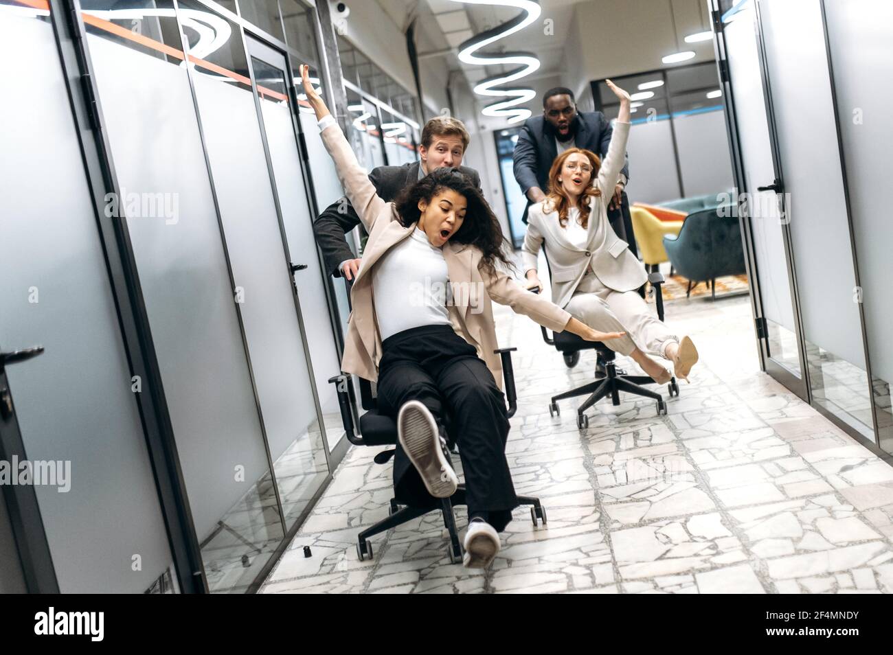 Funny young adult business people making a race on chairs in modern office.  Multiracial friendly colleagues spending together their work break, resting  from brainstorm Stock Photo - Alamy, image size:1300x954