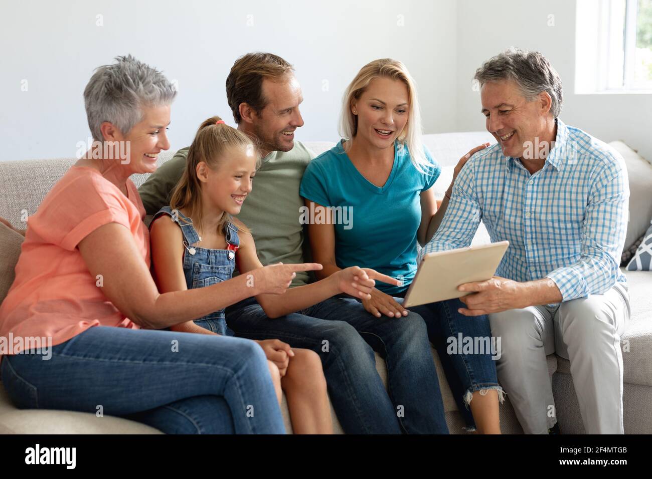 Smiling caucasian grandparents on couch with granddaughter and her parents looking at tablet Stock Photo