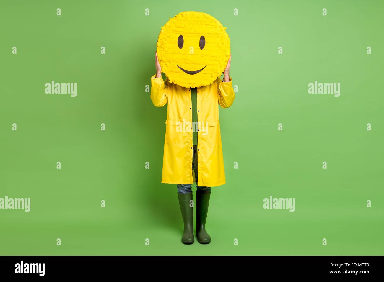Full body photo of young man close cover face paper pinata emoji smile wear raincoat isolated over green color background Stock Photo