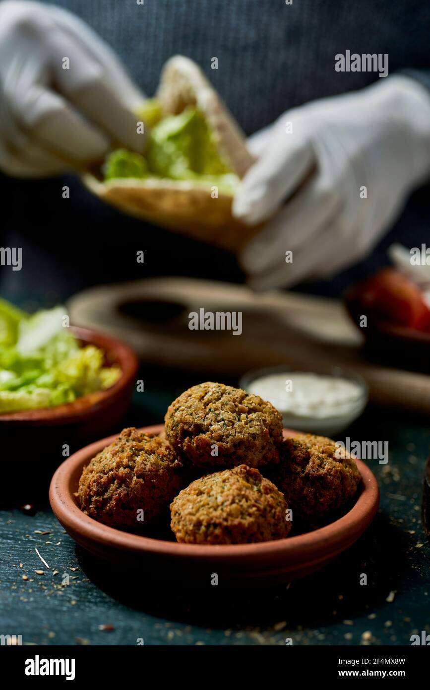 a young man, wearing latex gloves, prepares a falafel sandwich in a pita bread, filling it with chopped lettuce Stock Photo
