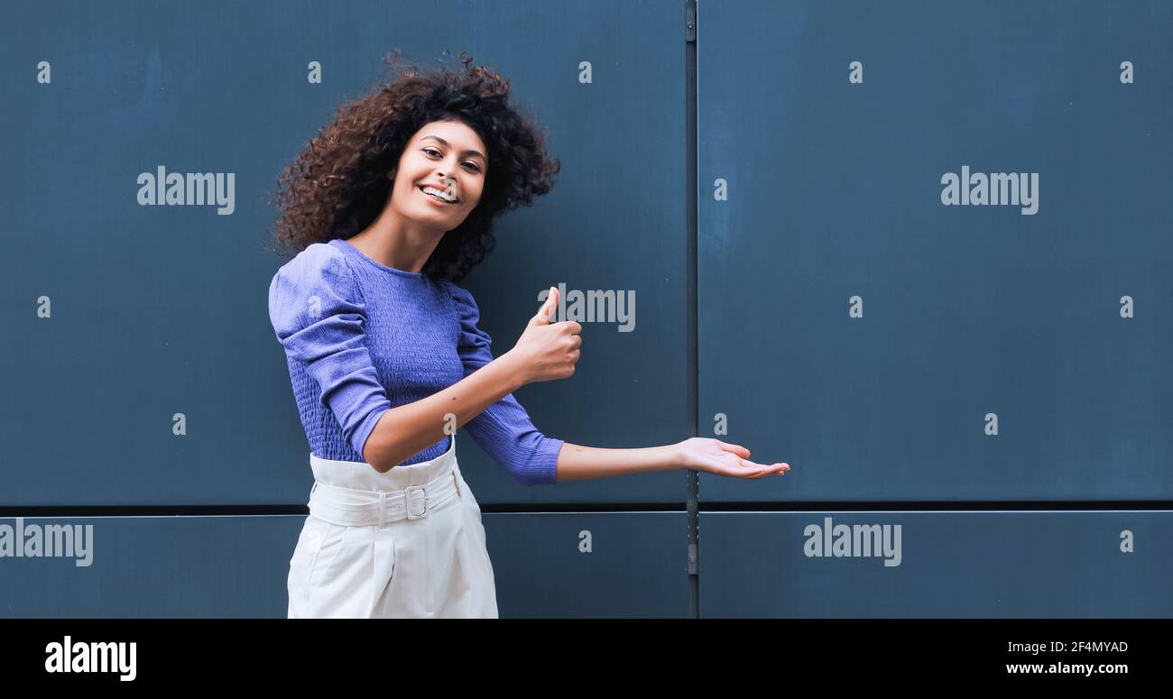cheerful young woman pointing with hand and showing thumb up outside Stock Photo