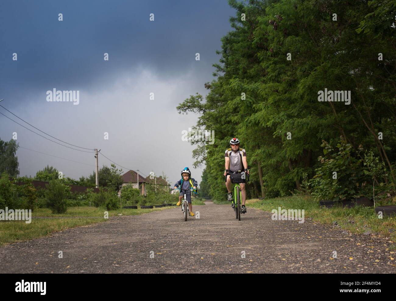 boy of 4 years old and man on bicycles are driving fast along road, in hurry to get away from thundercloud. Active healthy lifestyle. Be like dad. Fam Stock Photo
