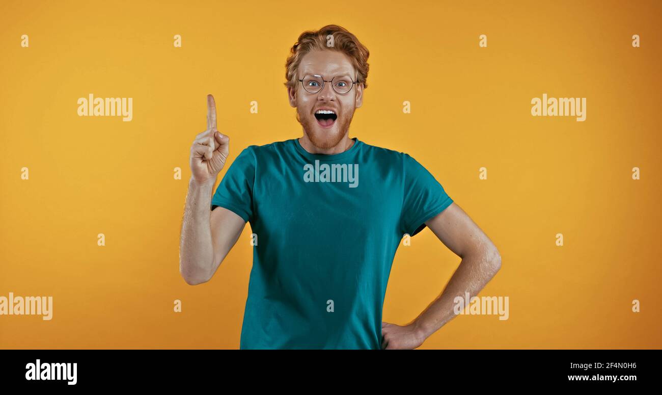 amazed redhead man in eye glasses standing with hand on hip while having idea isolated on yellow Stock Photo