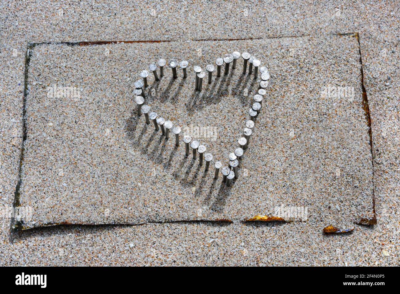 Heart of nails hammered into flattened tin over wooden plank, covered with beach sand. Stock Photo