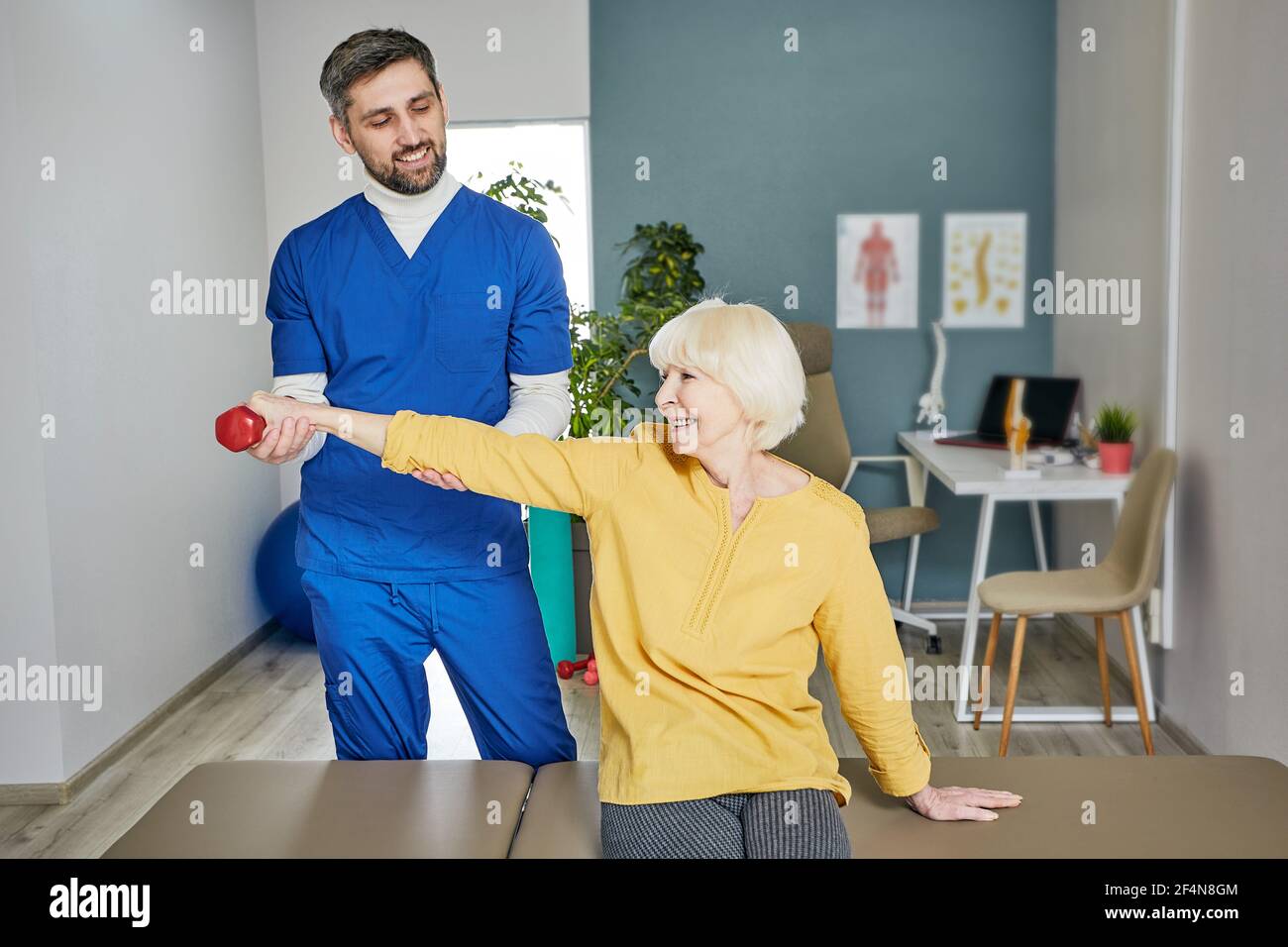 Senior woman lifts a dumbbell, doing treatment exercise with her physiotherapist. Physio