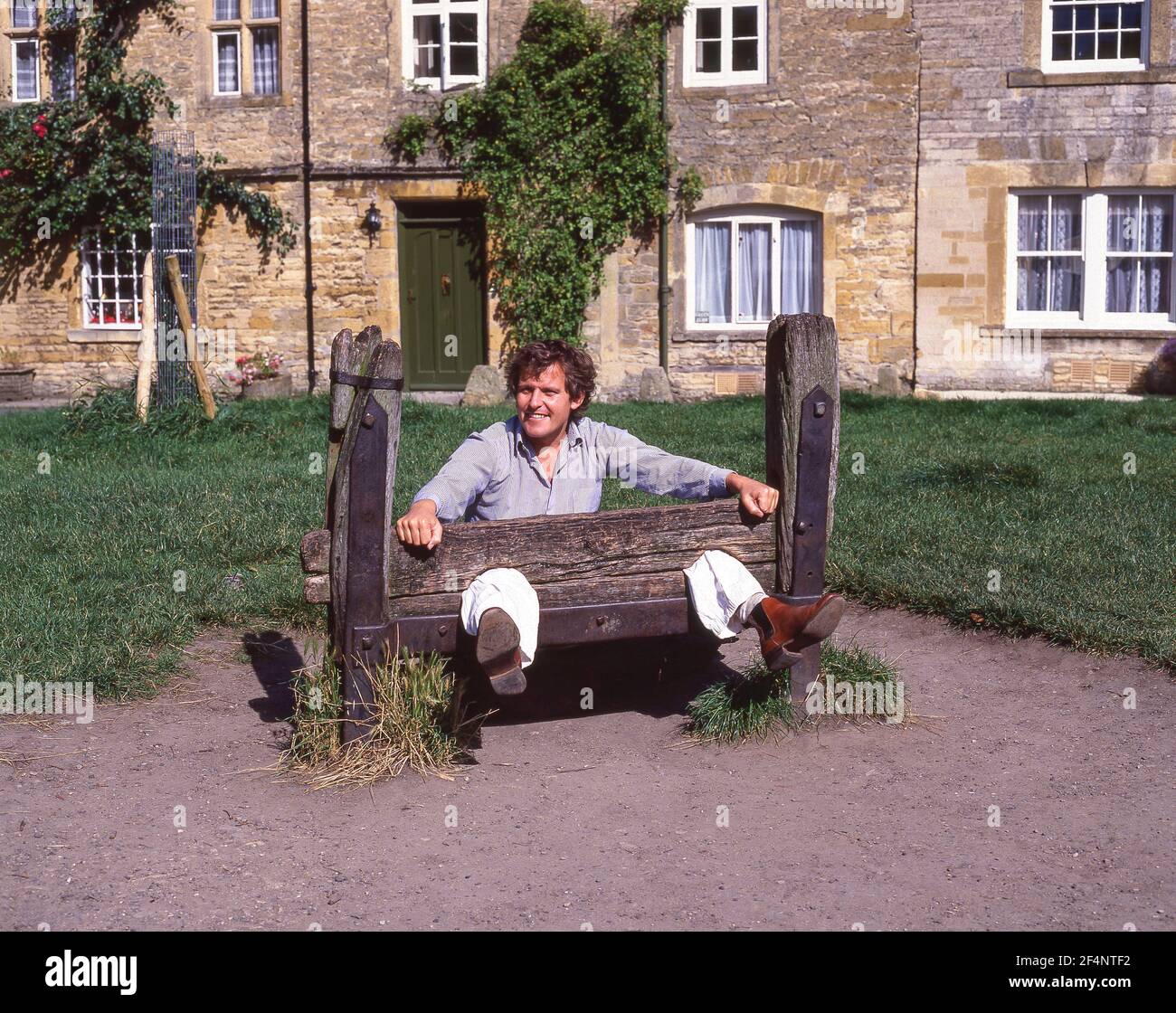 Young man posing in village stocks, Market Square, Stow-on-the-Wold, Gloucestershire, England, United Kingdom Stock Photo