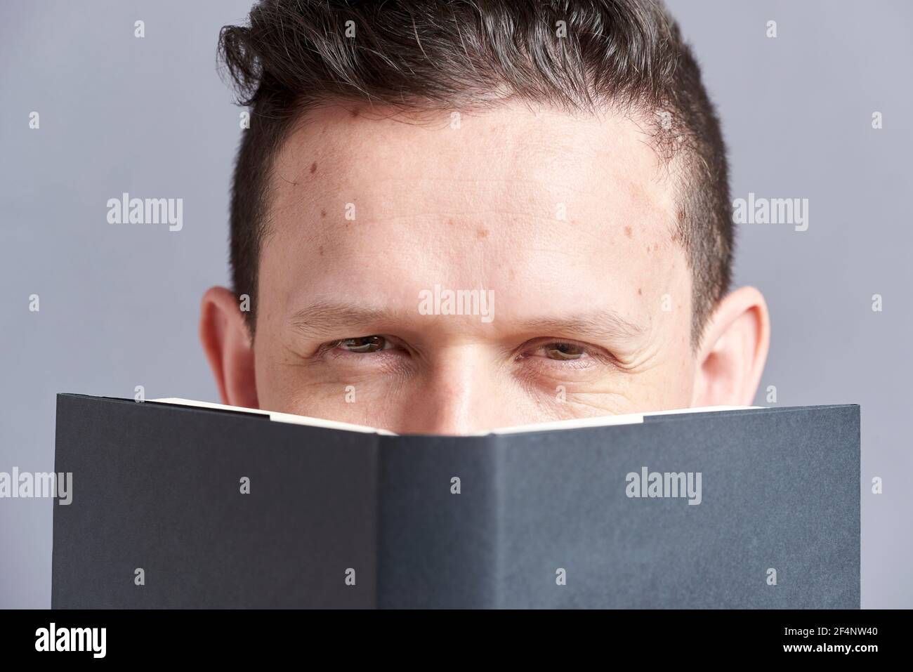 Young man looking intensely, askance, at the camera, peeking out from behind an open black book. Frontal portrait with gray background. Stock Photo