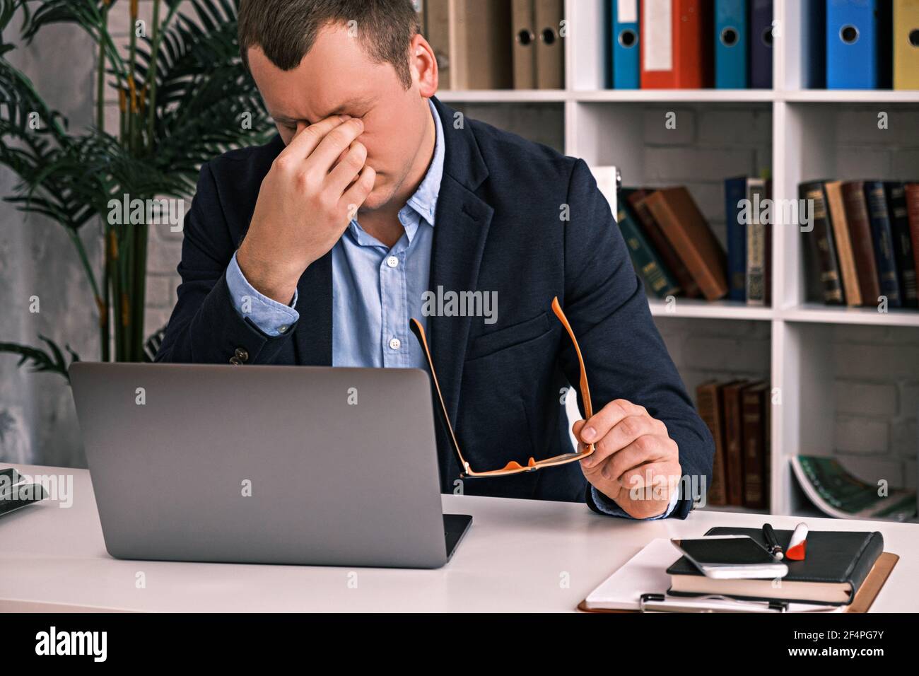 Eye fatigue, poor eyesight, office work. The young man takes off his glasses and rubs his eyes with his hand sitting at a table with a laptop in a sui Stock Photo