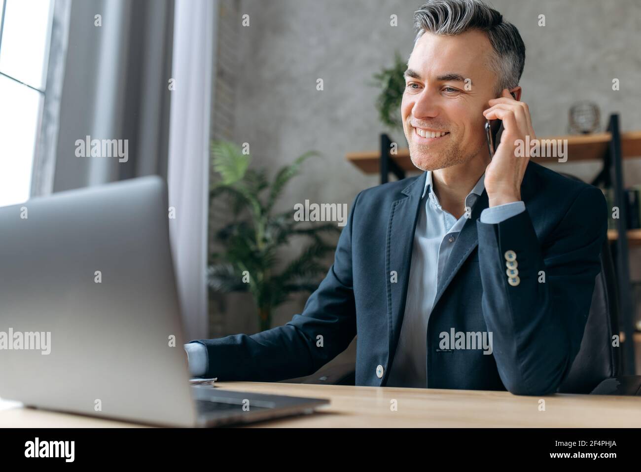 Busy attractive successful business leader sits at workplace in office, in a formal suit, works at a laptop, communicates with a client or employee by smartphone, smiles friendly looks at laptop Stock Photo