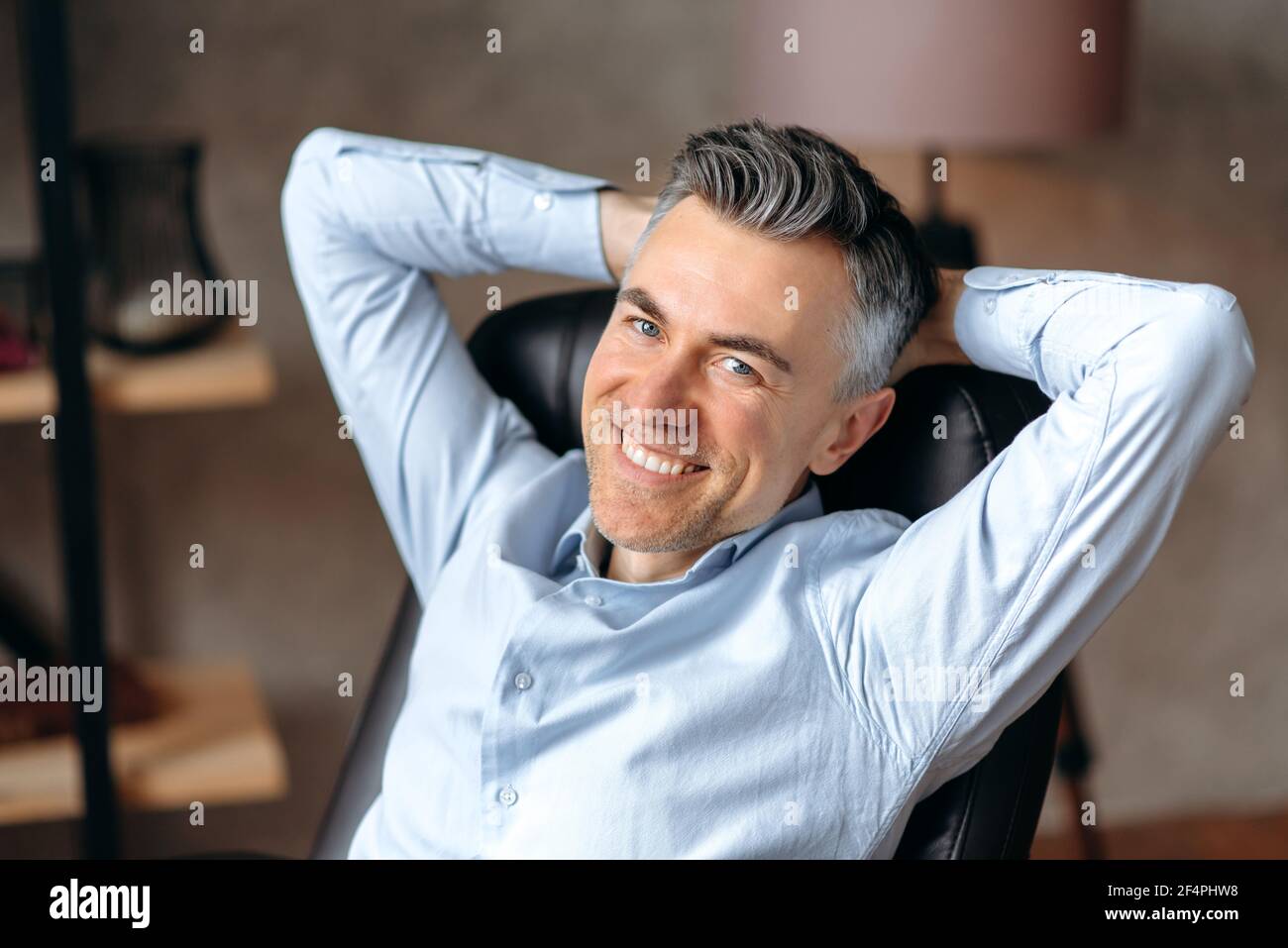 Portrait of a happy caucasian gray-haired man, manager or freelancer, sitting in modern office, wearing formal shirt, having rest from work, clasping his hands behind his head, smiling friendly Stock Photo