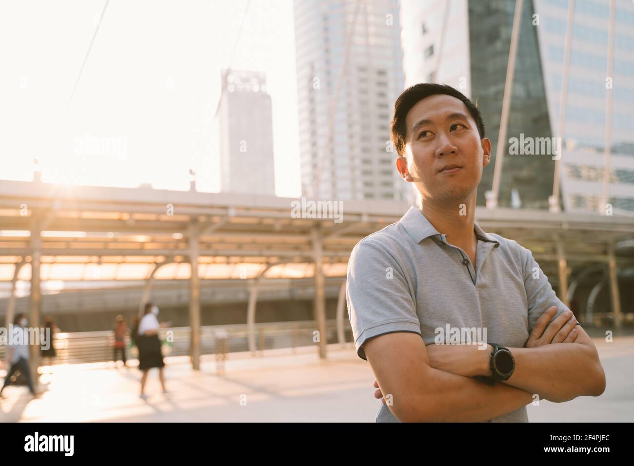 Portrait of unhappy young Asian man standing with arms crossed looking away while standing in city street worrying about future Stock Photo