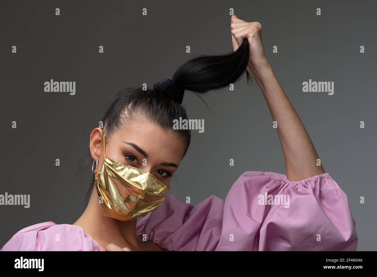 captivating look of a beautiful fashion girl touching her hair by twisting her ponytail; wearing a pink puff sleeve blouse and a gold surgical mask Stock Photo