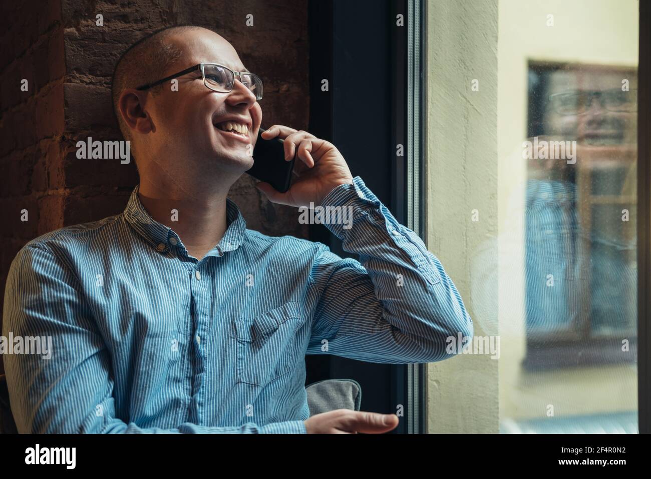 Man in a blue shirt sitting near the window and talking on smartphone. Working at home or modern coworking office. Stock Photo