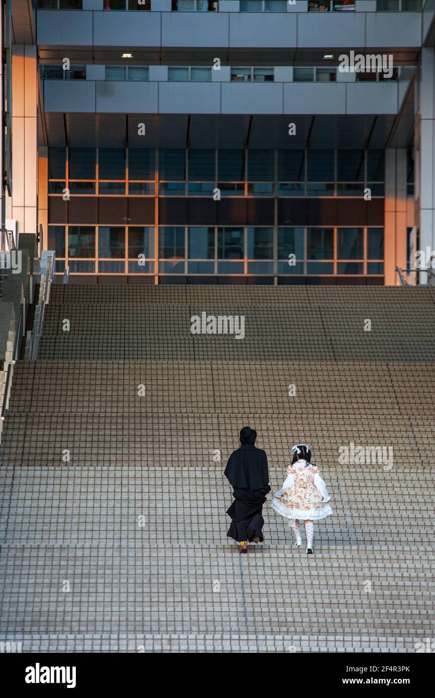 Distant rear view of Japanese couple: Sweet Lolita wearing pink dress, petticoat and male in frock coat climbing steep steps at Tokyo Bay, Japan Stock Photo