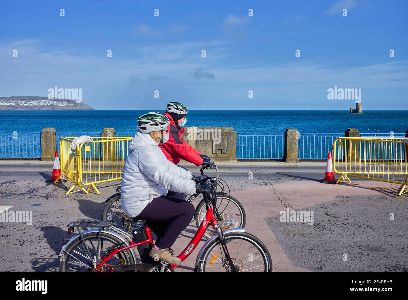 Two women cyclists using electric bikes riding on Douglas prom, Isle of Man wearing masks in Covid times Stock Photo