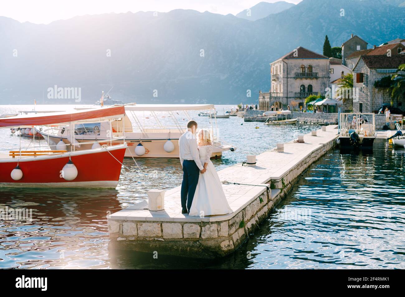 The bride and groom stand on the pier near Perast, holding hands and looking into each other's eyes, next to them boats  Stock Photo