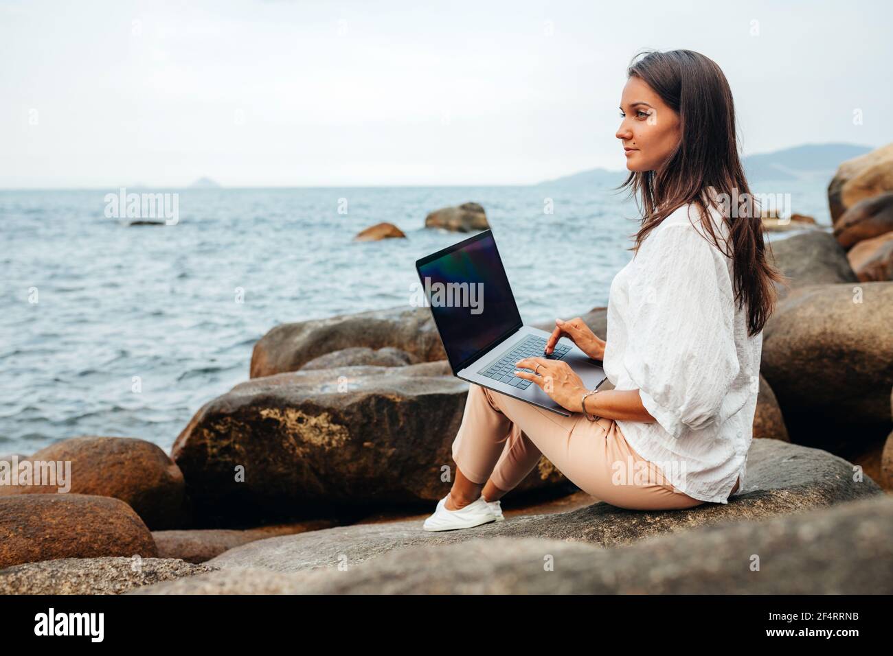 Freelance concept. A freelancer girl with a laptop on her lap works out of the office while sitting on a rock near the sea. Remote work in the fresh a Stock Photo