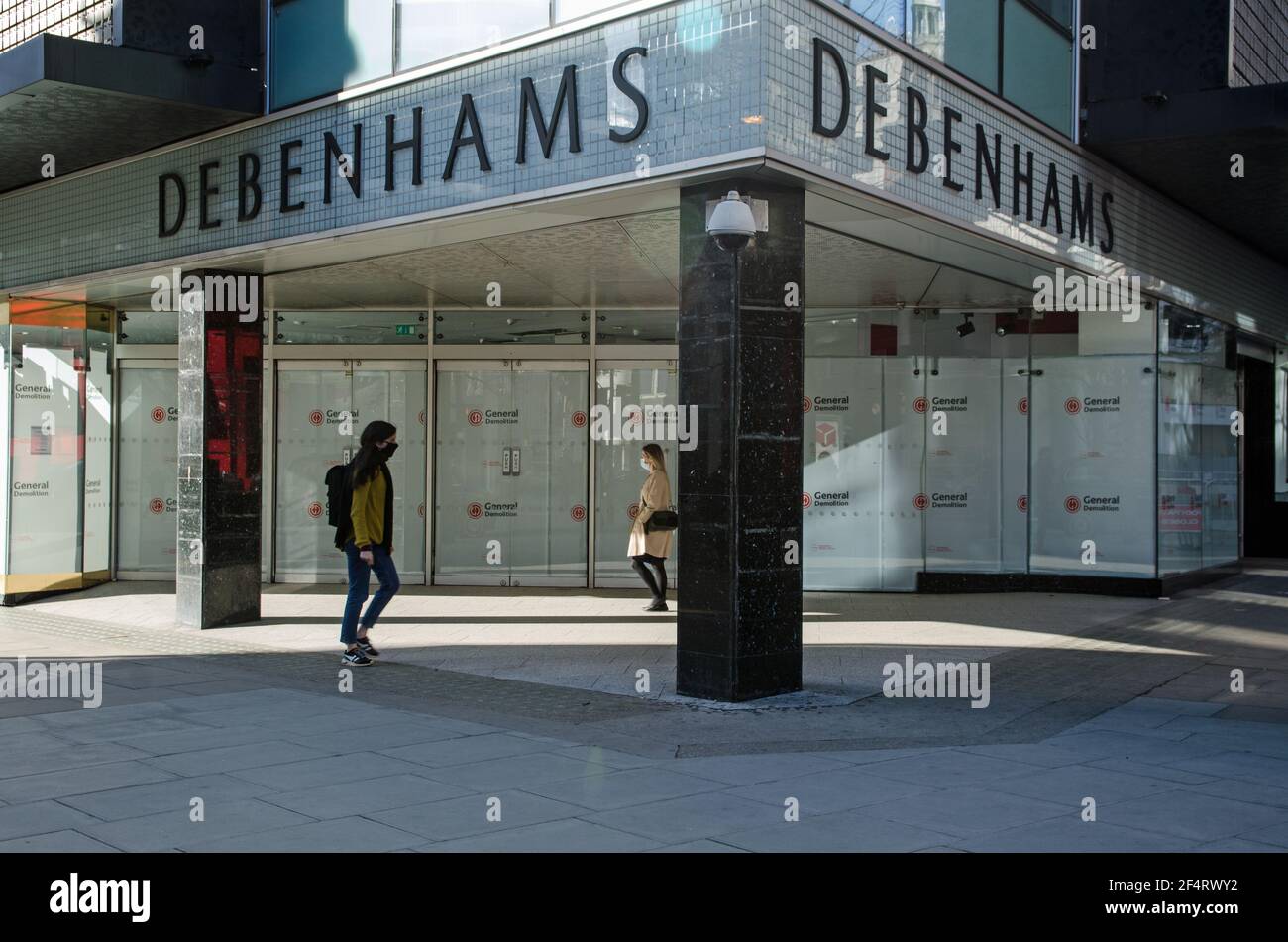 London, UK - February 26, 2021: Two pedestrians, each wearing face masks walking past the closed doors of the now defunct flagship store of the Debenh Stock Photo
