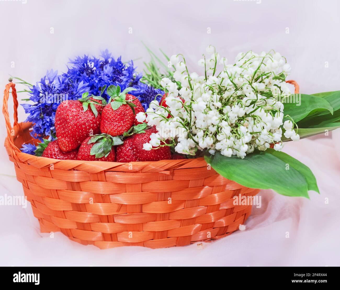 Pretty lilies of the valley, red fresh strawberries and cornflowers in orange basket Stock Photo
