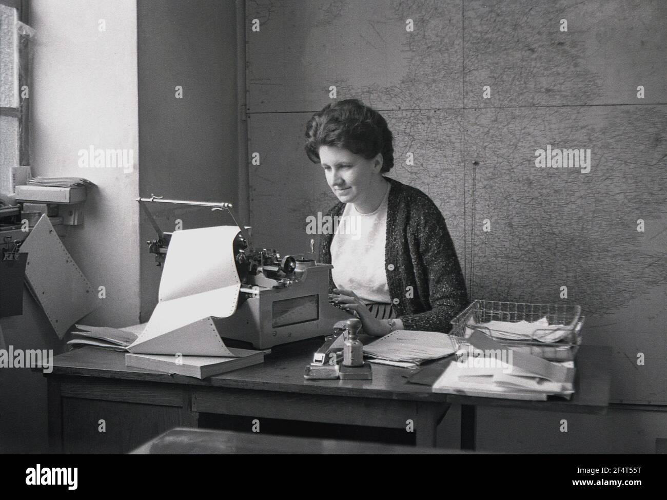 1964, historical, inside an office at a trading company on an industrial estate, a lady administrator sitting at a wooden desk using a typewriter, England, UK. A large map of Britain displayed on the wall behind her. Stock Photo