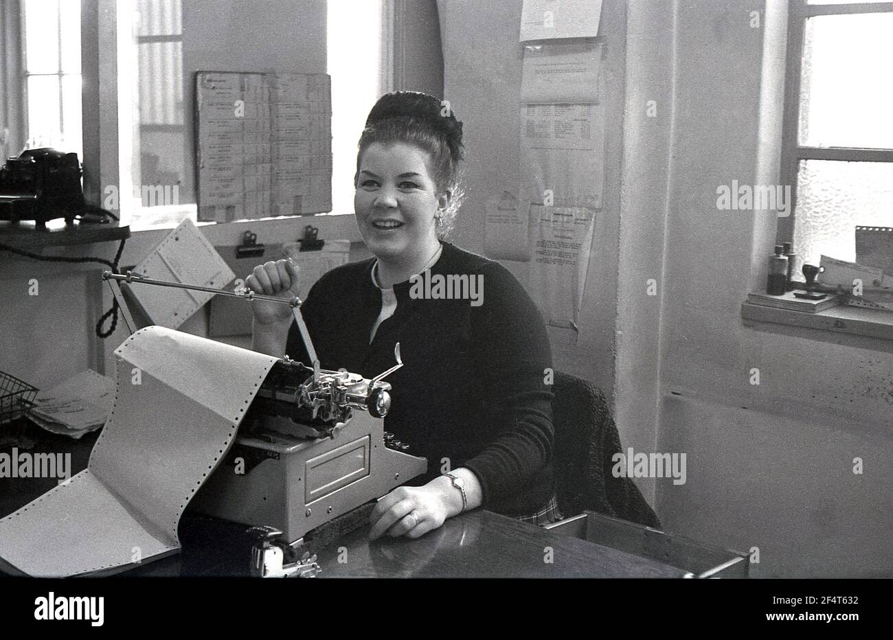 1964, historical, inside an office of a trading company selling rotavators and cultivators, a female clerical worker sitting at a wooden desk at a typewriter, with a large print out in, England, UK. Stock Photo