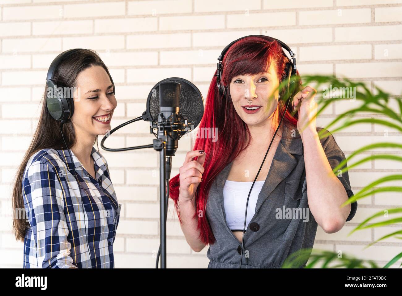 Happy young women with headset standing near microphone and enjoying singing against brick wall and plant Stock Photo