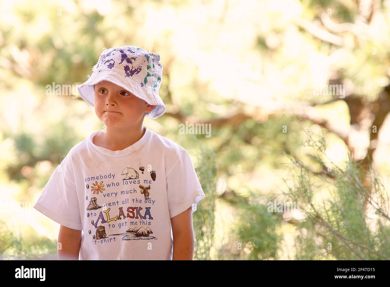 Puzzled boy in t shirt and hat pressing lips together and looking away on blurred background of countryside on sunny summer day Stock Photo