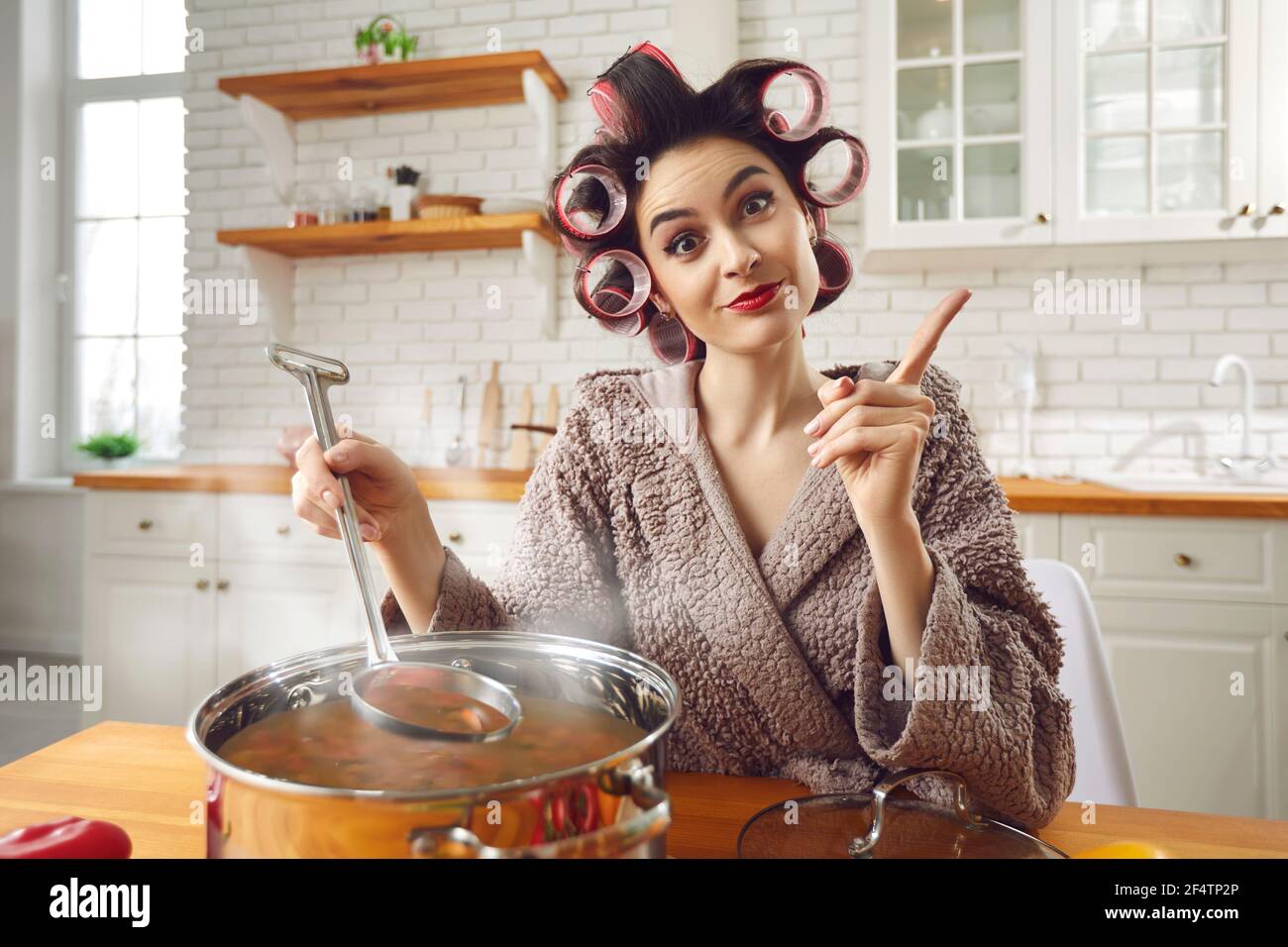 Young funny attractive housewife in curlers and bathrobe cooking at home kitchen Stock Photo
