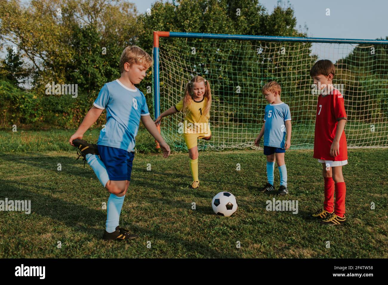 Children exercising together outside at football goal. Group of kids in soccer dresses stretching together at amateur competition in field at sunset. Stock Photo