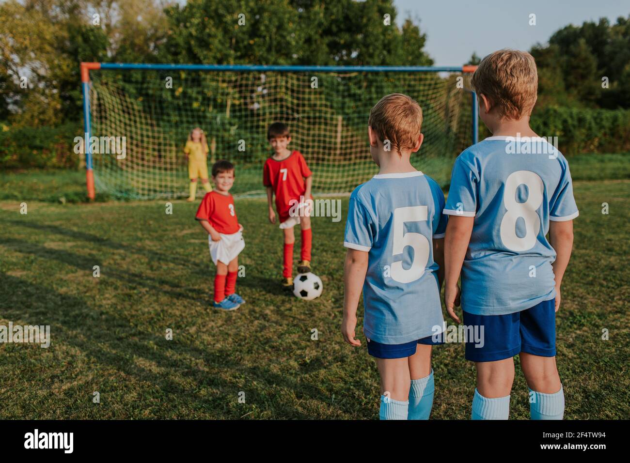 Children playing amateur football. Kids in teams wearing soccer dresses playing soccer in field at sunset. Stock Photo