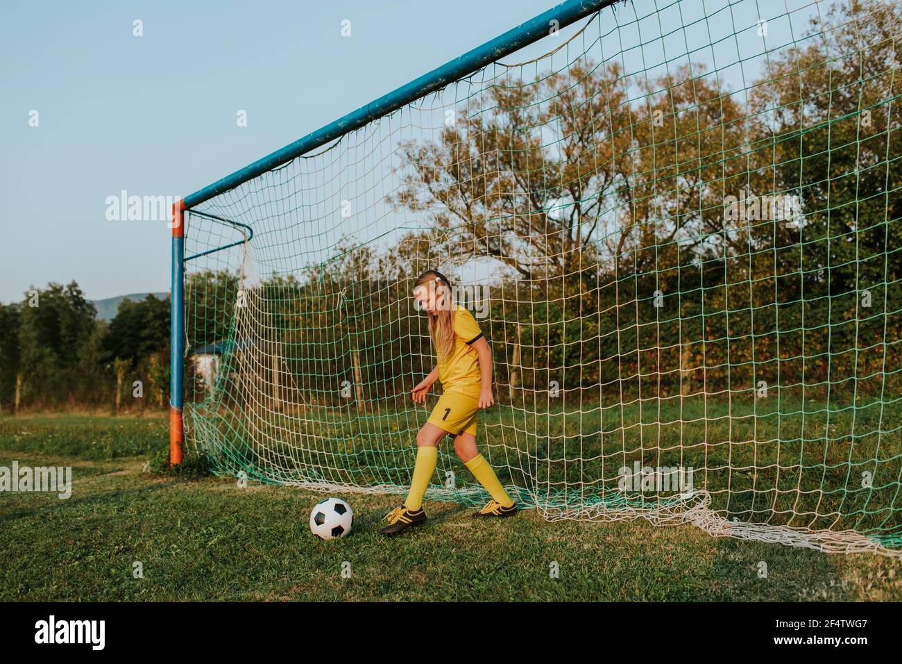 Girl goalkeeper defending goal. Young girl footballer in yellow football dress kicking ball out of net. Stock Photo