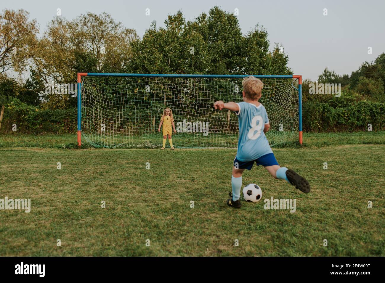 Back view of a child wearing football dress shooting ball in front of goal. Full length of girl goalkeeper standing at goal and soccer player kicking Stock Photo