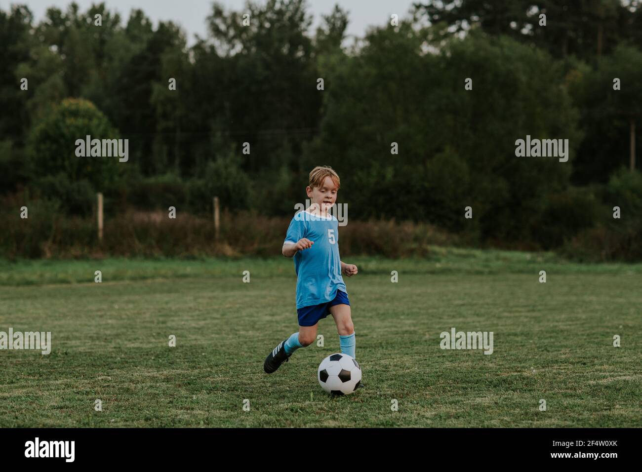Child soccer player with ball. Boy in blue football dress running after ball on field outside. Stock Photo