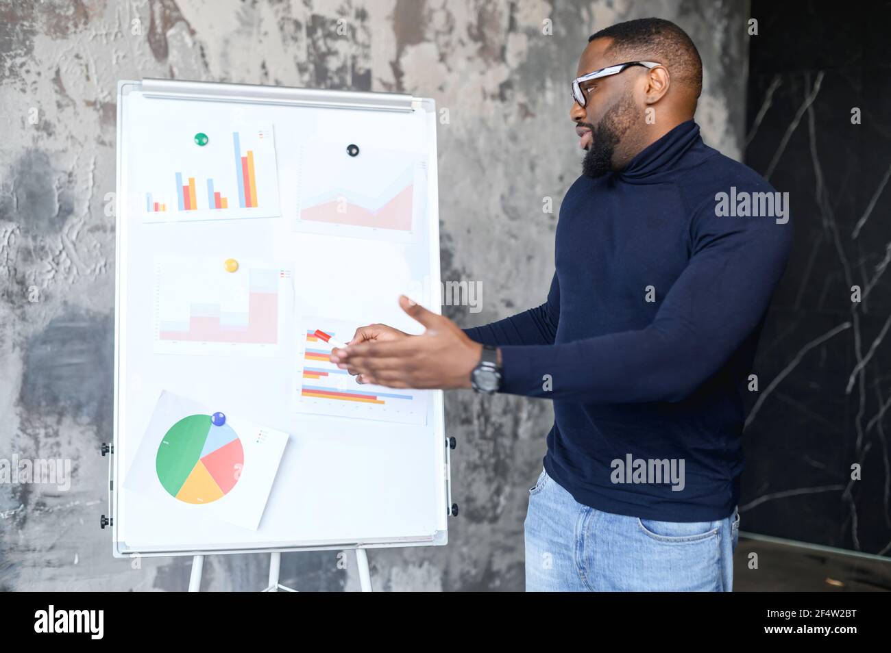 Confident African-American male employee points on whiteboard with graphs, planning strategy, smart and intelligent freelancer guy presenting new project, sturtup idea, office employee reports Stock Photo