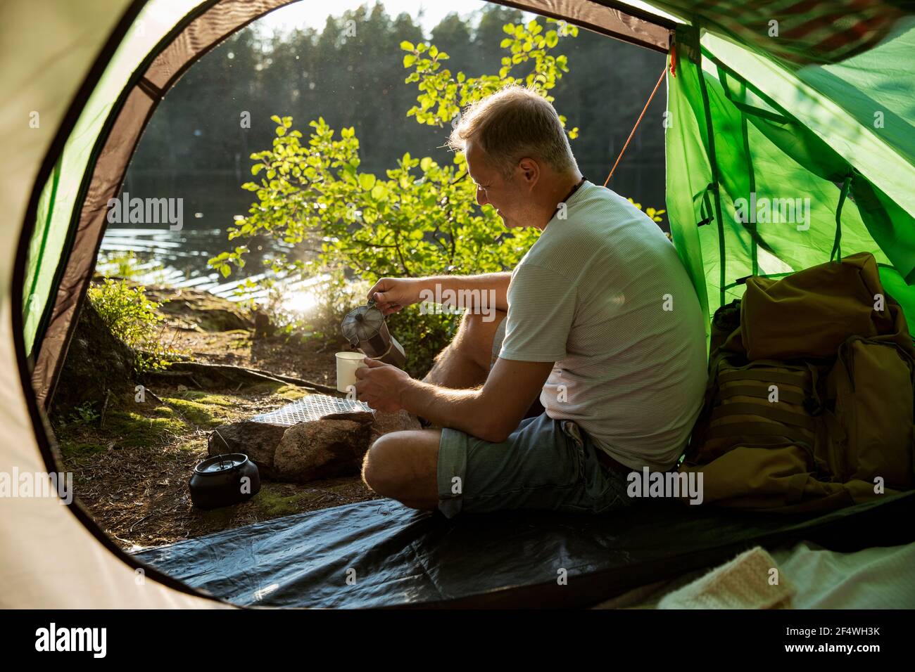 Man making coffee using espresso maker on campfire in forest on shore of a lake, sitting in tent, making a fire, grilling. Happy isolation concept. Stock Photo