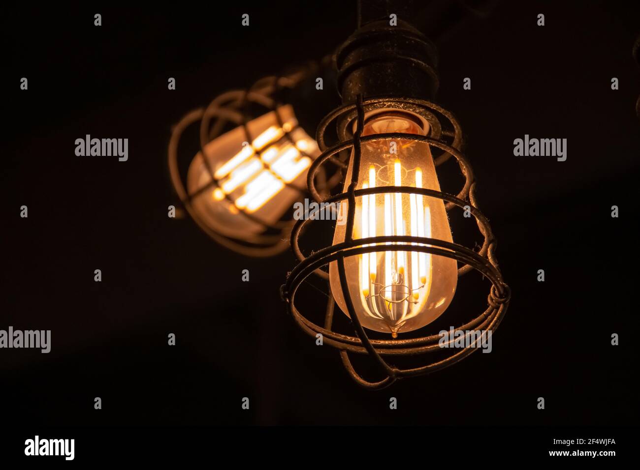 A red-hot factory incandescent light bulb with glowing filaments inside of an iron cage hanging from the ceiling of a pub in London, Ontario, Canada, Stock Photo
