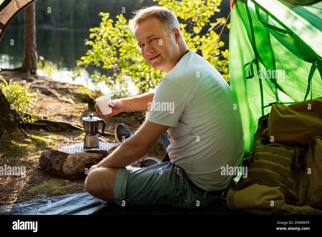 Man making coffee using espresso maker on campfire in forest on shore of a lake, sitting in tent, making a fire, grilling. Happy isolation concept. Stock Photo