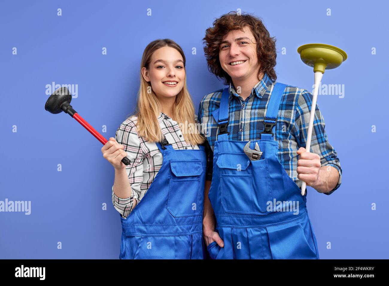 Positive team of two plumbers dressed in blue uniform with plunger, being glad to repair bathtub, recieve pleasant comments from clients, isolated ove Stock Photo