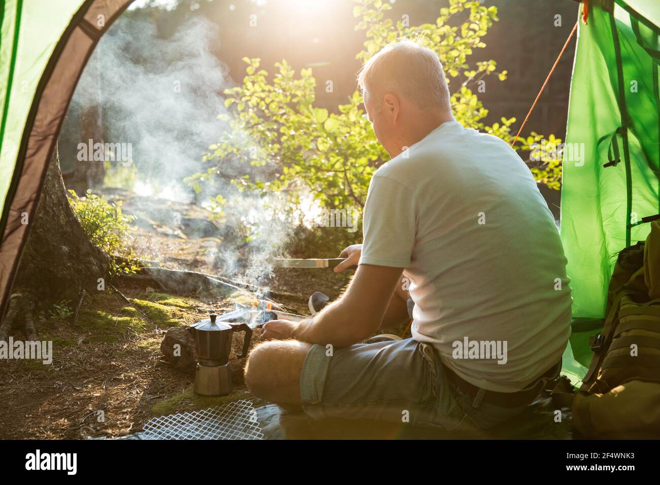 Man making coffee using espresso maker on campfire in forest on shore of a lake, sitting in tent, making a fire, grilling. Happy isolation concept. Stock Photo