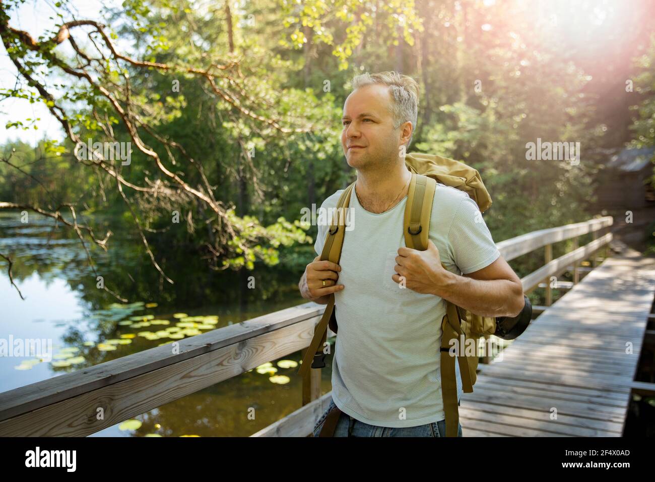 Mature man exploring Finnish nature in summer, walking across the bridge. Hiker with big backpack traveling in forests. Summer Scandinavian landscape Stock Photo