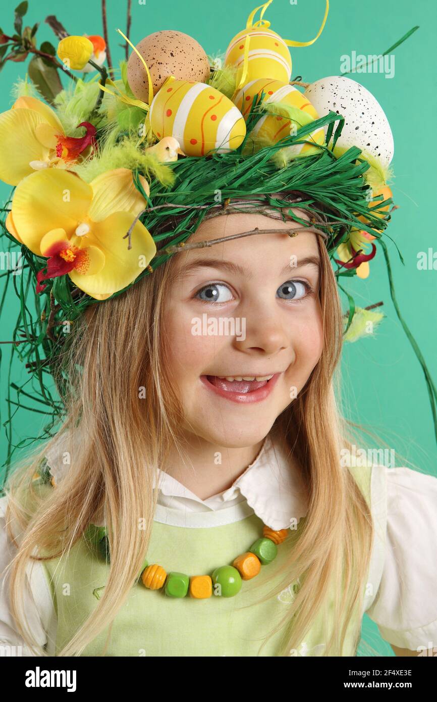 Little smiling blonde girl wearing a costume of yellow Easter eggs, spring flowers and bird feathers on green background. The child looks at the camer - Stock Image