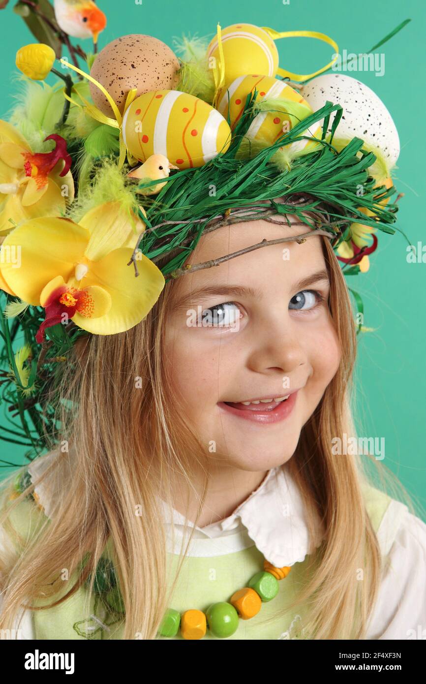Portrait of a smiling blonde girl wearing hair dressing of yellow Easter eggs, spring flowers and bird feathers on a green background. The child looks - Stock Image