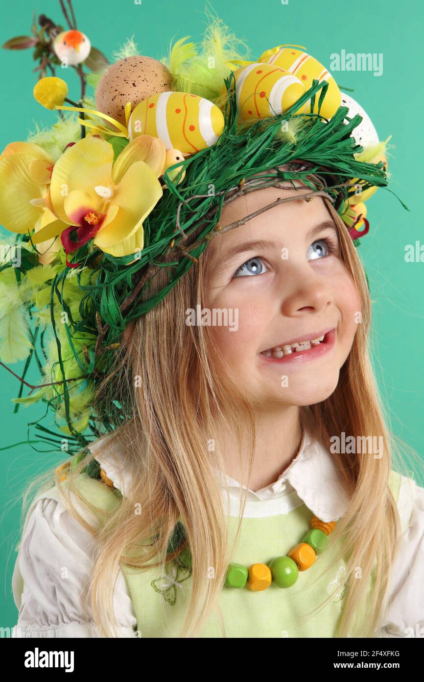 Happy Easter day. seven year old girl with festive Easter hairstyle made of yellow easter eggs, spring flowers and bird feathers. Smiling child looks - Stock Image