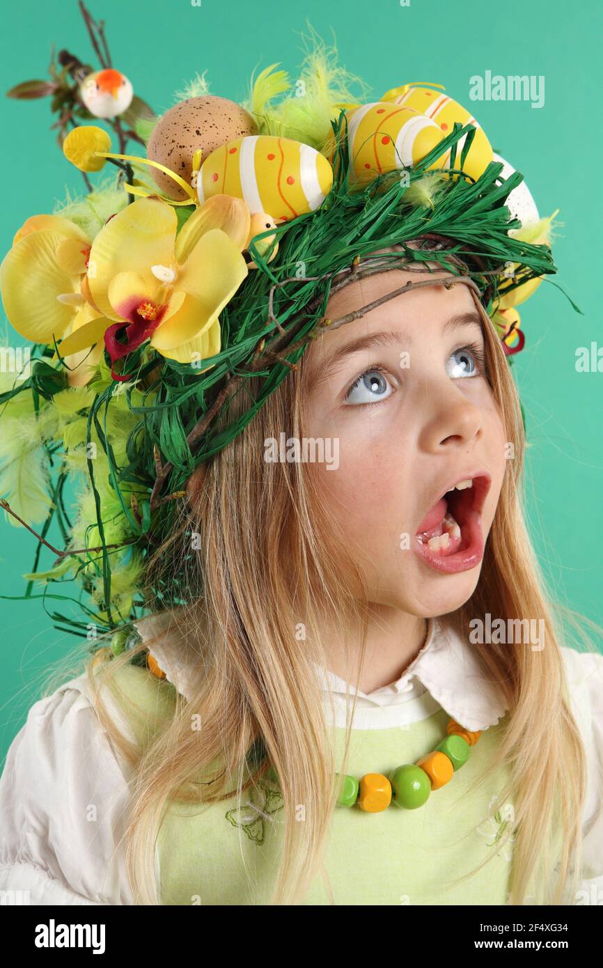 Cute little girl dressed in a straw hat with yellow Easter eggs, spring flowers, feathers and birds. The child looks upwards by opening his mouth - Stock Image
