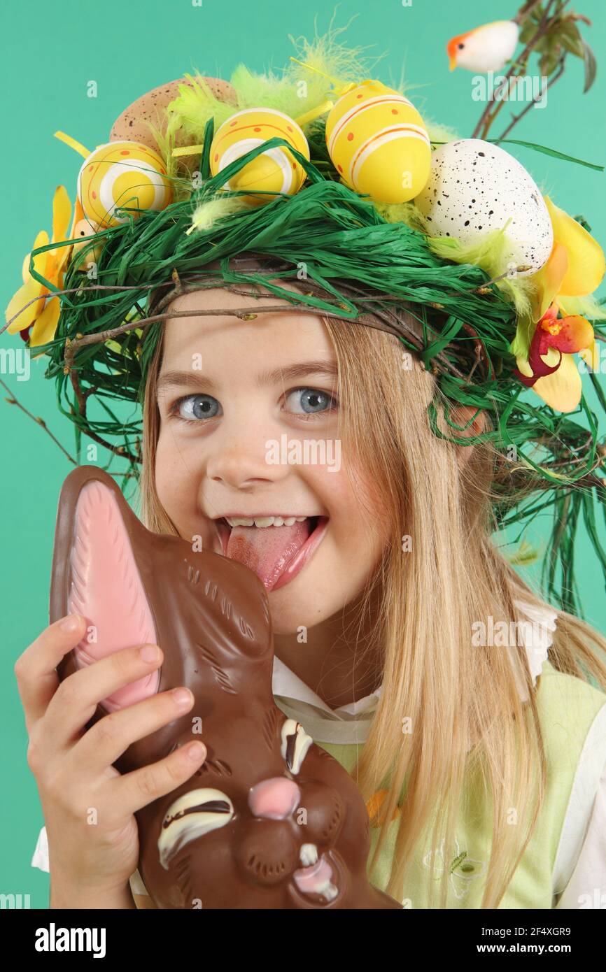 Easter concept. Child eating chocolate bunny. The young Girl wears a festive headdress made of yellow Easter eggs, spring flowers and bird feathers. Stock Photo