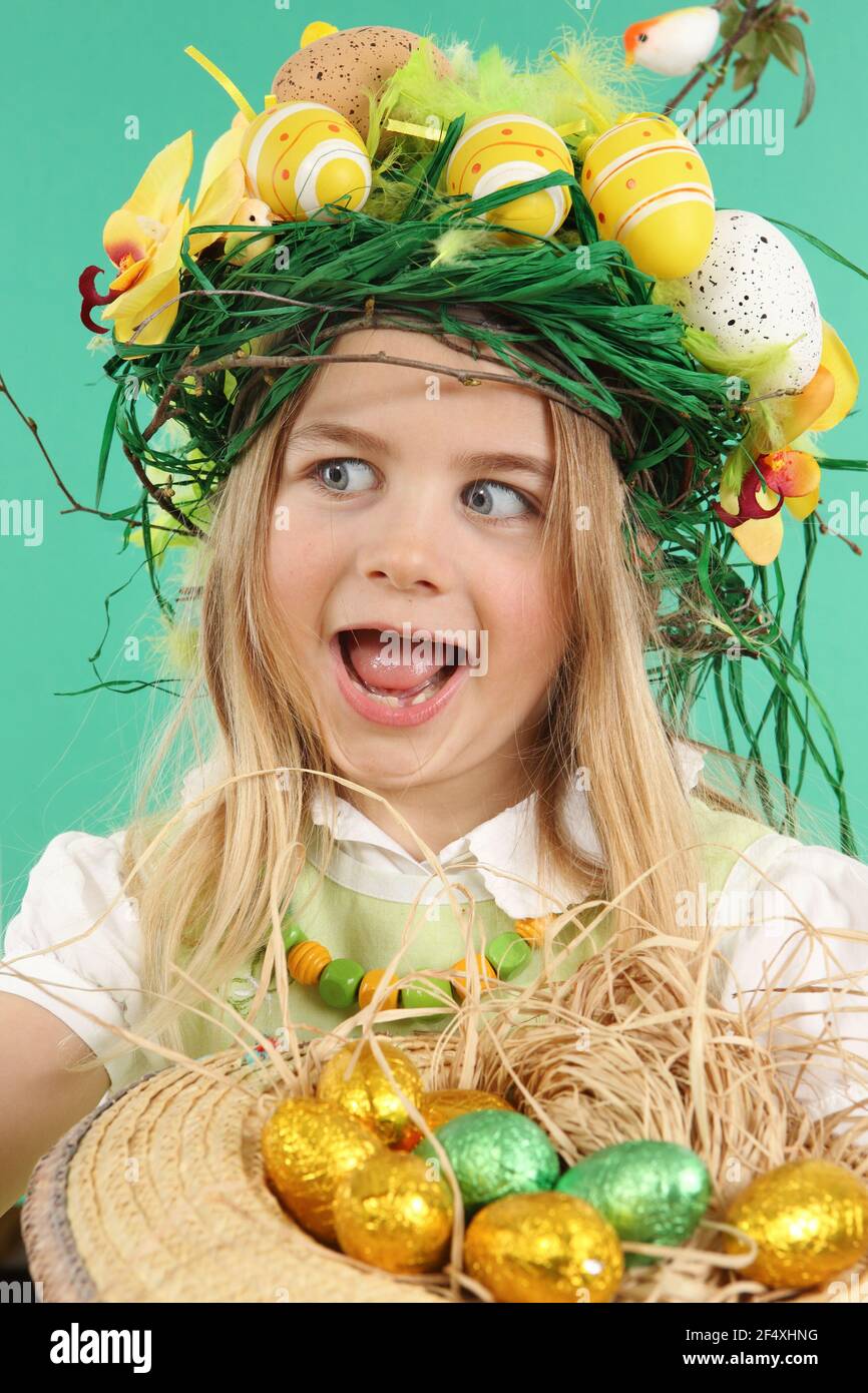Child disguised with a basket of Easter eggs, looks to the side. The girl wears a festive headdress made of yellow Easter eggs, spring flowers and bir - Stock Image