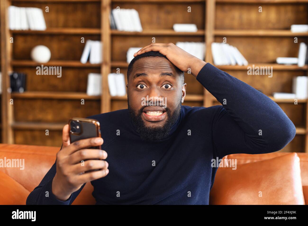 Discouraged African-American man grabbed his head, holding a smartphone, looks at the camera with eyes wide opened, portrait of shocked black guy with phone in hand Stock Photo
